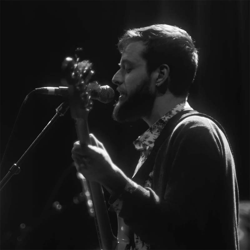 White male singing into a microphone while playing a guitar, in black and white.