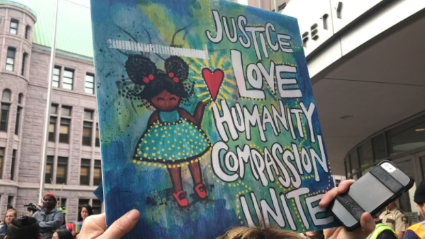 Protest sign reading "Justice, Love, Humanistyh, Compassion, Unite" with a drawing of a Black girl with puffs holding a heart. She is wearing a blue and yellow polka dot dress and red Mary Jane shoes.