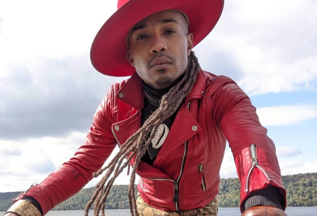 Je wears a bright red leather jacket and brimmed hat. He has blonde dreads and light brown skin